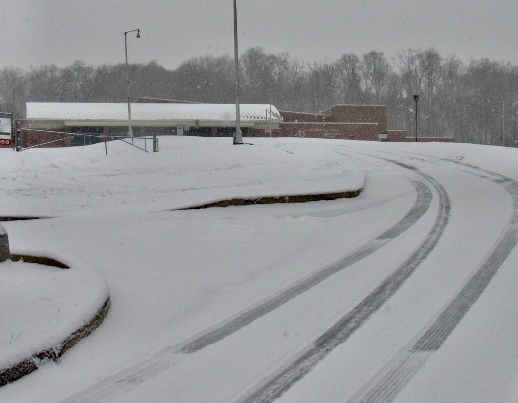 A snowy scene showing a snow-covered driveway leading to a school building. The building, identified as the Columbia Borough School District’s Hill Campus, is partially visible in the background with trees lining the horizon. Snow continues to fall, and tire tracks are visible on the curved road in the foreground. A streetlight and sign are present along the driveway.